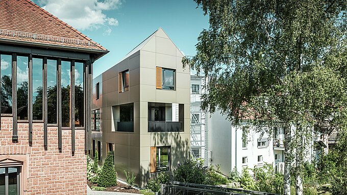 The image shows the modern town hall in Alzenau, whose façade is made of bronze-coloured aluminium composite panels from PREFA. The pointed roof and clearly structured windows with wooden blinds give the building an elegant and contemporary aesthetic. In the foreground is a green area with a paved path and trees, which harmoniously complements the urban design of the building.