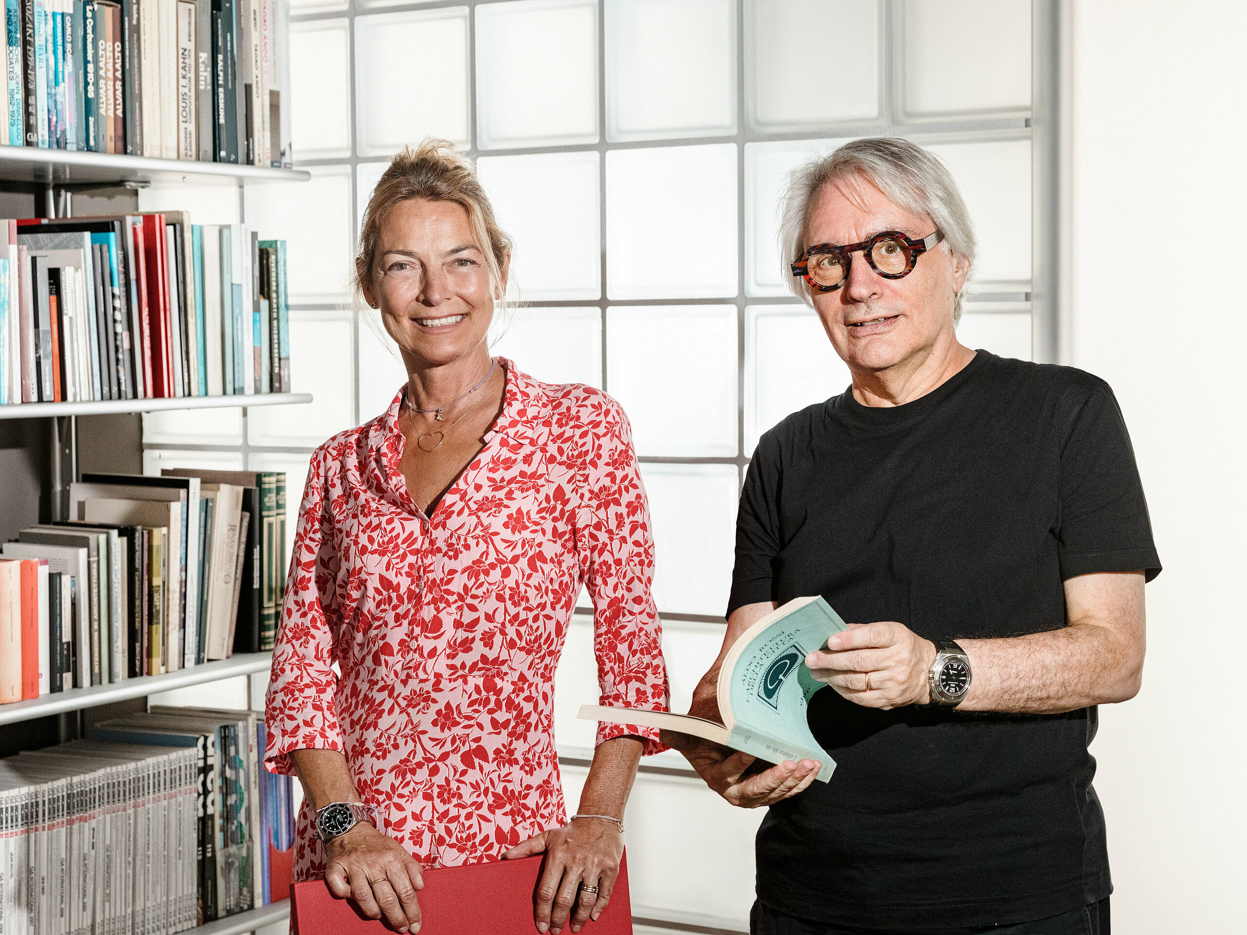 Bolzano architect Claudio Lucchin and his office partner Daniela Varnier pose in front of a modern glass block wall that refracts the light in an exciting way. Claudio Lucchin is dressed entirely in black and holds a book in his hands. Daniela Varnier wears a bright red top and smiles warmly. This stylish setting symbolises innovation and transparency. The scene represents forward-looking architecture, as realised in the ‘Tempesti’ barracks in Corvara in South Tyrol.