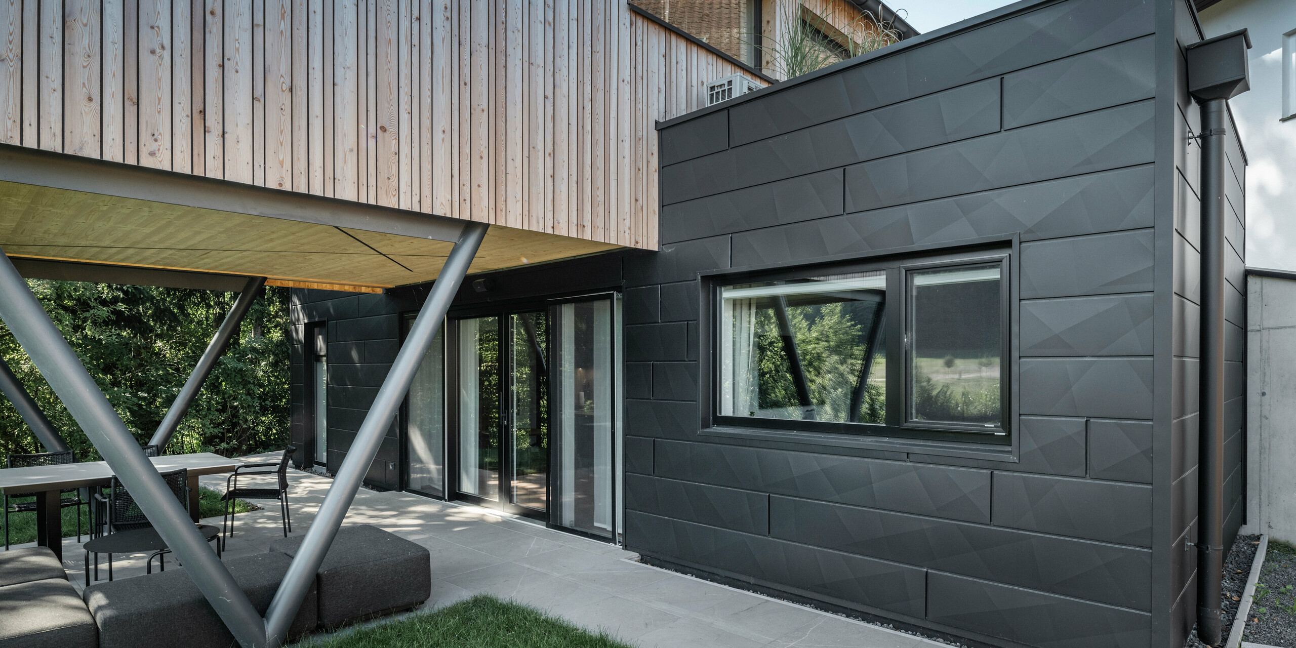 Architectural highlight: “Woodstock Chalet 5” in St. Martin am Innkreis, Upper Austria. The modern wedding venue is accentuated by the use of anthracite-coloured PREFA Siding.X façade panels. The aluminium elements form a striking contrast to the light-coloured wood panels and the metallic support structures. The dark window frames blend harmoniously into the façade design, giving the building an elegant appearance. This image shows the aesthetic balance between natural wood texture and the industrial charm of the aluminium façade, which is representative of contemporary building design.