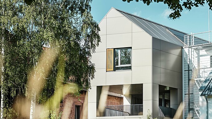 The image shows the modern town hall in Alzenau, whose façade is made of aluminium composite panels from PREFA in an elegant bronze tone. The pointed roof area and large windows emphasise the contemporary character of the building. In the foreground, trees and green spaces frame the building, while the soft lighting and clear sky underline the modern urban space.
