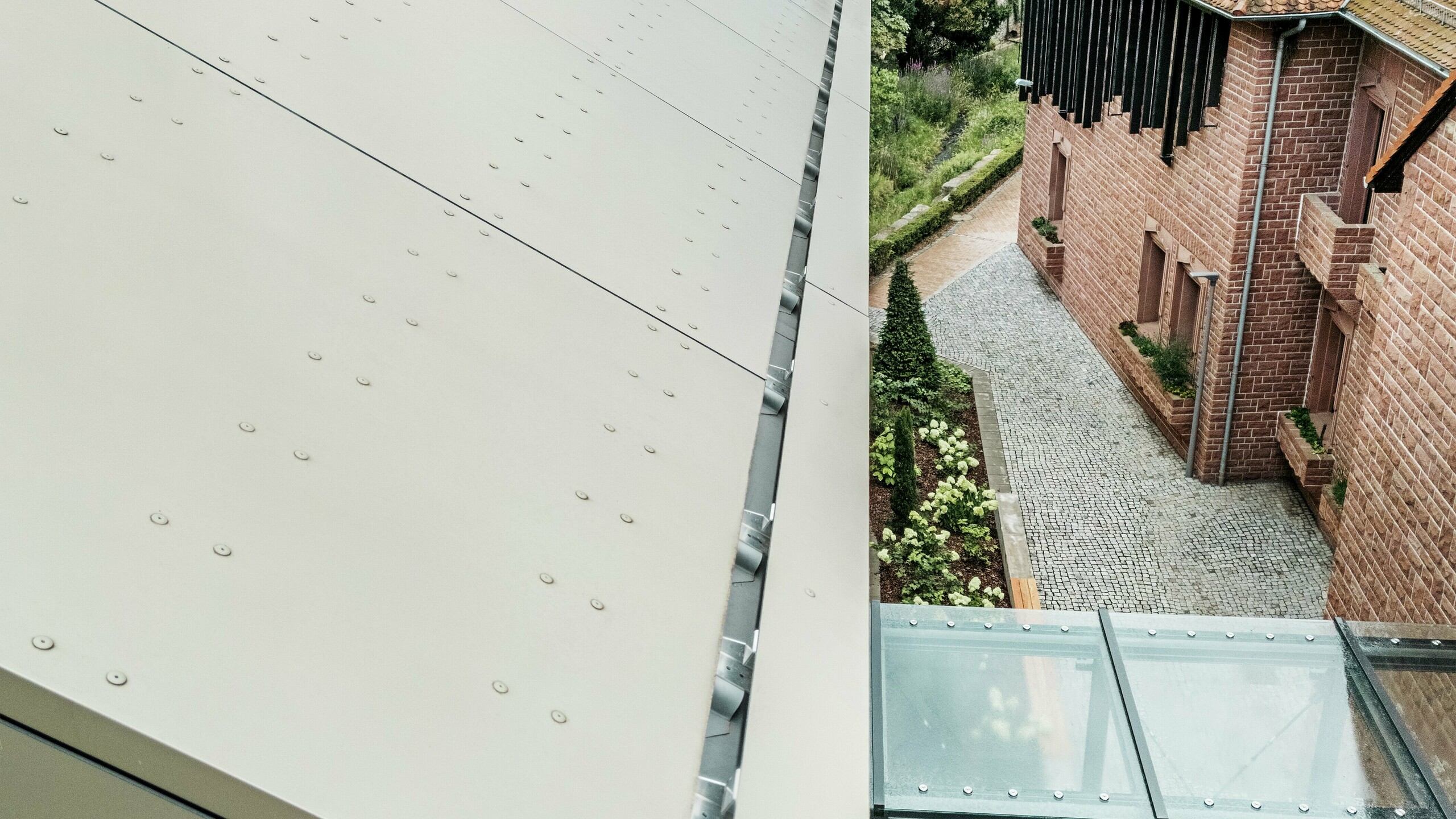 Close-up of the roof of the town hall in Alzenau, which is covered with PREFA aluminium composite panels in a soft bronze tone. The panels are precisely laid and fitted with visible fasteners, giving the façade a modern, industrial look. In the foreground, a glass walkway can be seen connecting the town hall to an adjacent building.