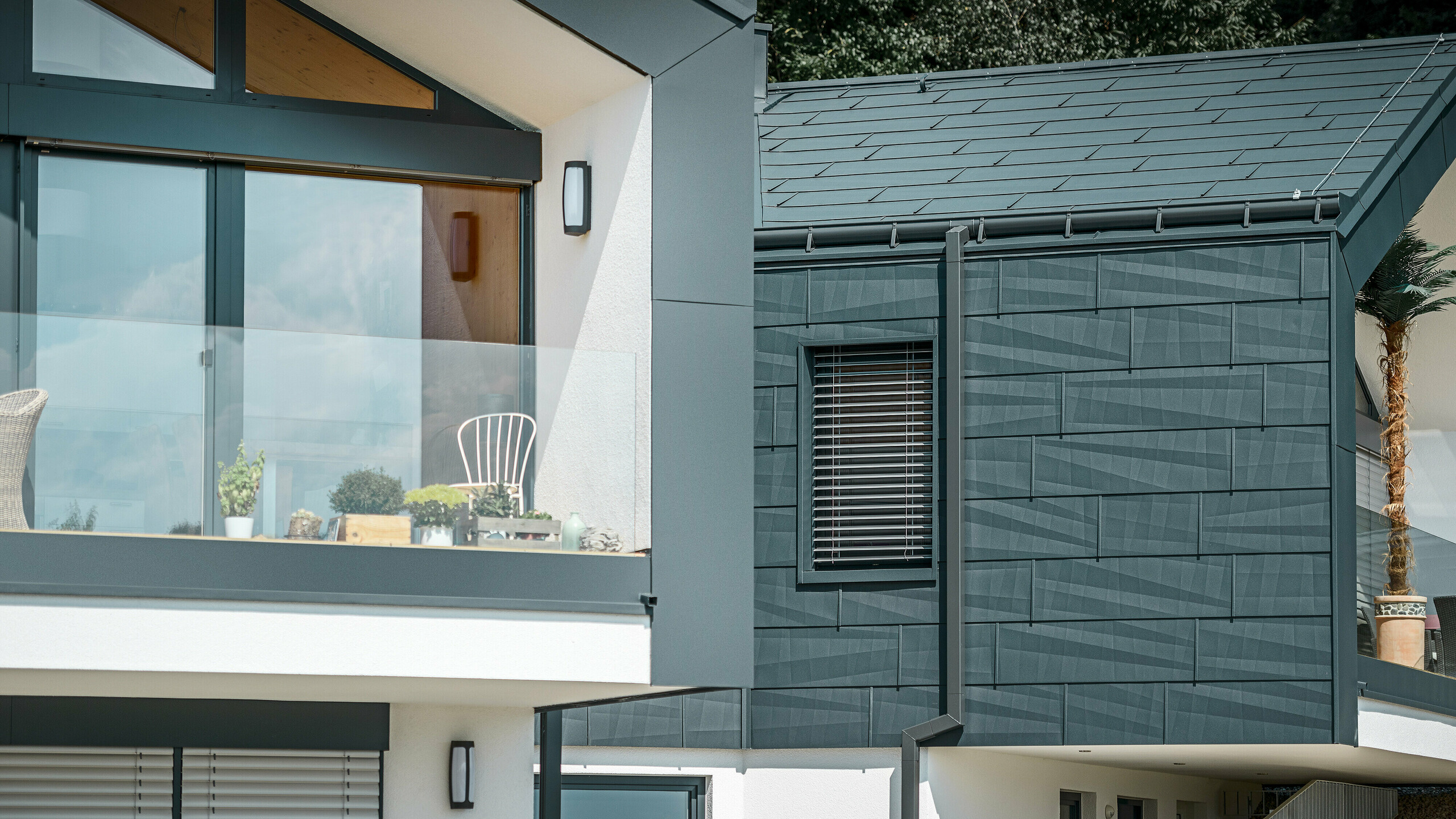 The image shows a modern semi-detached house with a stylish aluminium façade. The façade is finished in a dark anthracite colour and has a textured surface thanks to PREFA's FX.12 roof and façade panels. A on-roof gutter and square downpipe from PREFA have been installed on the roof as a drainage system. A balcony with glass railings can be seen on the upper floor, while windows with slatted blinds are found on the lower floor. The architecture appears clear and minimalist, and trees can be seen in the background, creating a natural atmosphere.