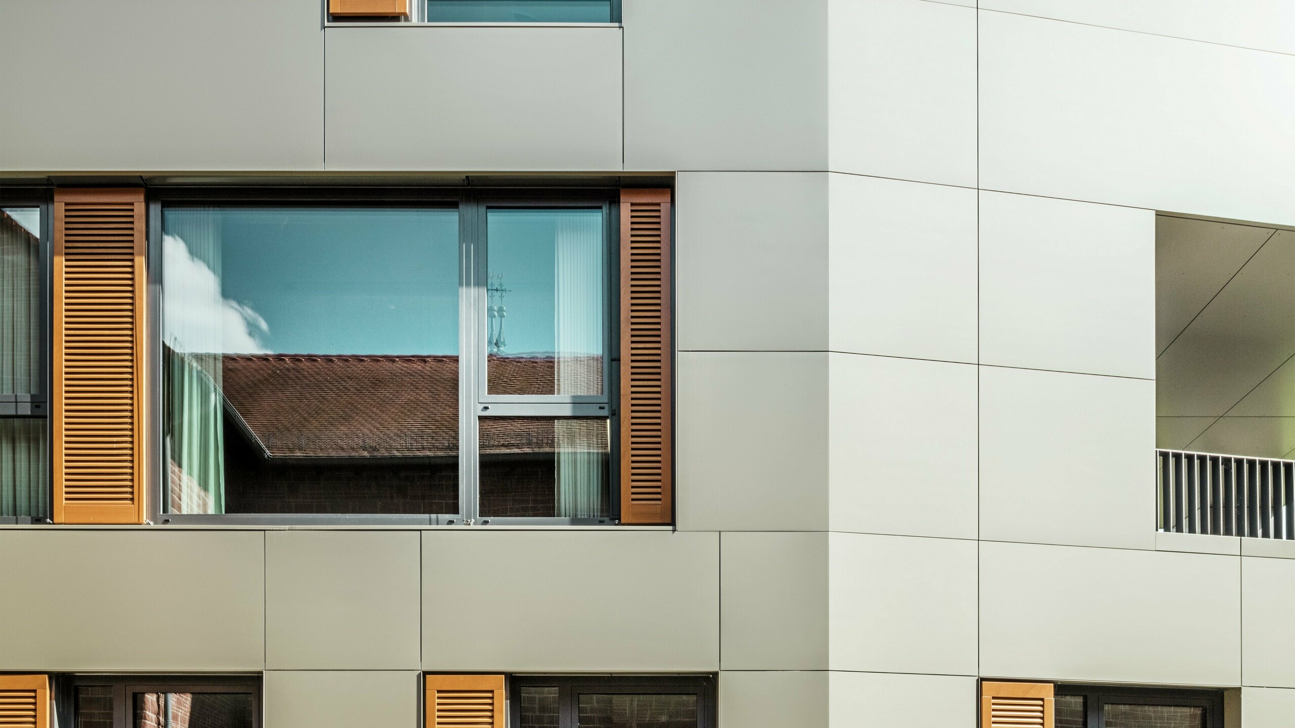 Close-up of the façade of the town hall in Alzenau, Germany, with aluminium composite panels from PREFA in the colour bronze. The large windows are framed by elegant wooden blinds in a warm brown, which give the façade an attractive texture. In the foreground, a well-tended flower bed complements the urban design of the building with green accents. The clear sky and clean lines of the architecture emphasise the modern character of the building.
