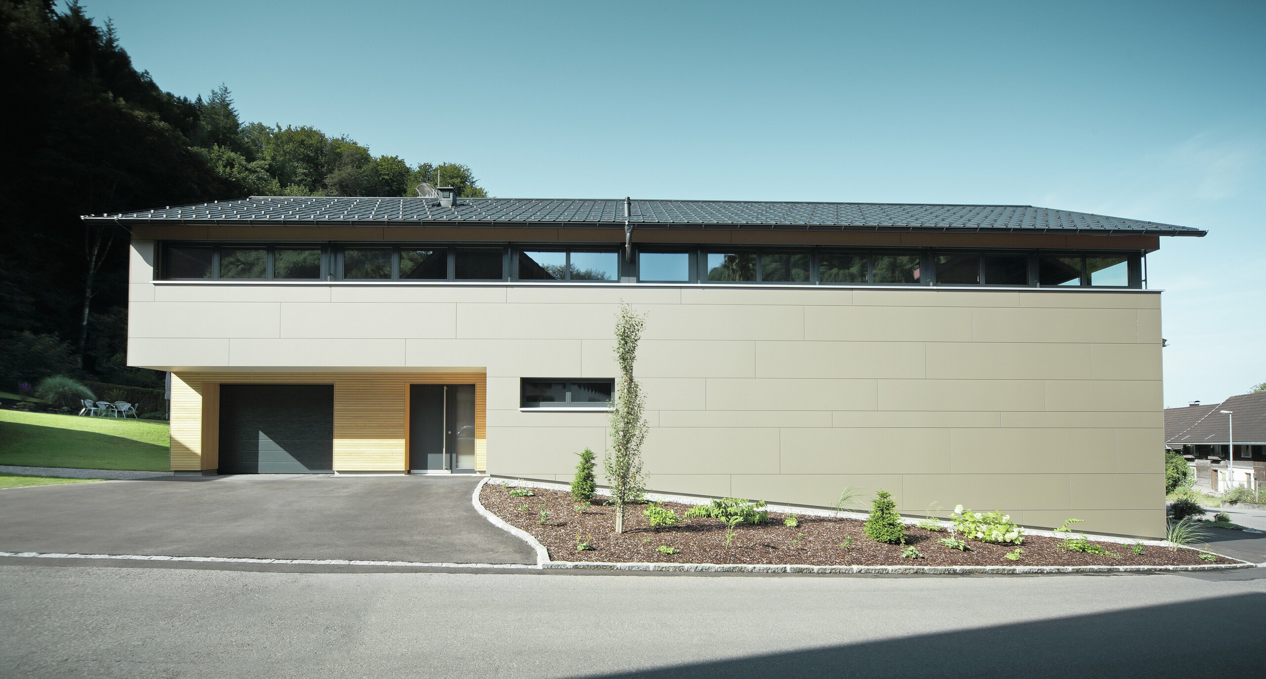 Das Einfamilienhaus in Hohenems präsentiert sich in einer modernen und stilvollen Fassade aus PREFA Aluminium Verbundplatten in Bronzemetallic, mit einem darüberliegenden Dach aus PREFA Dachplatten in P.10 Anthrazit. Die klare Linienführung und das durchdachte Farbkonzept des Designs integrieren das Gebäude nahtlos in die natürliche Umgebung. Die Gestaltung zeigt die Kombination aus Funktionalität und Ästhetik, die PREFA Produkte für zeitgemäße Wohnarchitektur bieten.