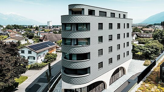 Luftaufnahme des Bernerhauses in Buchs mit markanter, dreieckiger Kubatur und weich gerundeten Balkonen. Die Fassade aus PREFA Wandschindeln in P.10 Hellgrau umschließt das Gebäude wie eine schuppenartige Aluminiumhaut und sorgt für ein lebendiges Spiel aus Licht und Schatten. Über dem massiven Betonsockel mit eleganten Arkaden wirkt die leichte, witterungsbeständige PREFA Aluminiumfassade besonders präzise, langlebig und wartungsarm – ein moderner Eyecatcher inmitten der alpinen Stadtlandschaft.