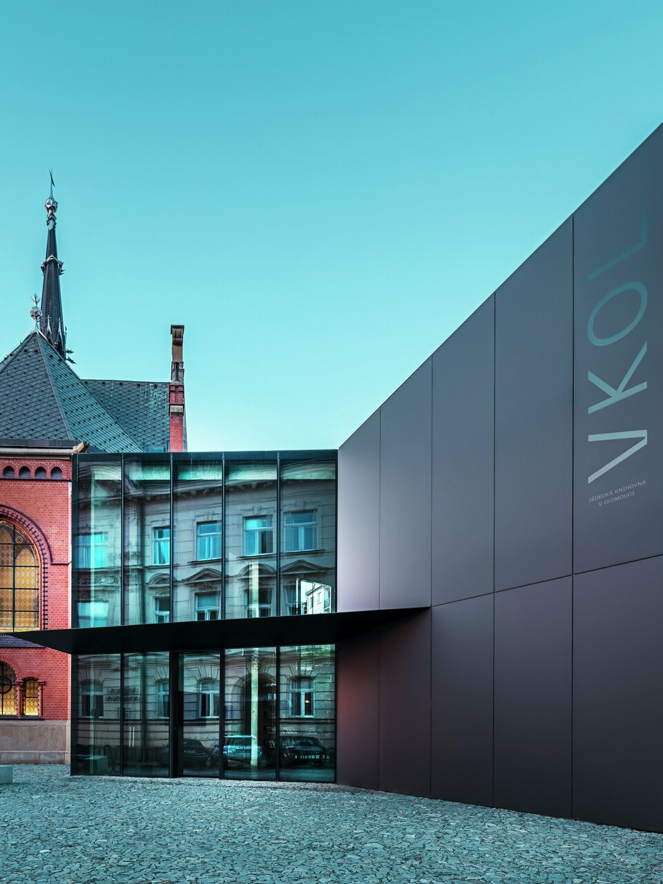 Das Bild zeigt das imposante Zusammenspiel zwischen der historischen Roten Kirche und dem modernen Neubau in Olmütz. Die Kirche mit ihren charakteristischen gotischen Fenstern und dem markanten Turm bildet das historische Rückgrat des Ensembles, während der moderne Anbau mit seiner eleganten, schwarzen PREFABOND Aluminium-Verbundplatten-Fassade eine zeitgemäße, architektonische Antwort darstellt. Große Glasflächen lassen den Blick in das Innere des Neubaus zu, wo die Umgebung durch Reflexionen in den Fenstern eine interessante Wechselwirkung zwischen Alt und Neu widerspiegelt. Die präzise Verarbeitung der PREFABOND-Platten betont die moderne Ästhetik, während die harmonische Integration in das historische Umfeld sowohl funktional als auch visuell beeindruckt.