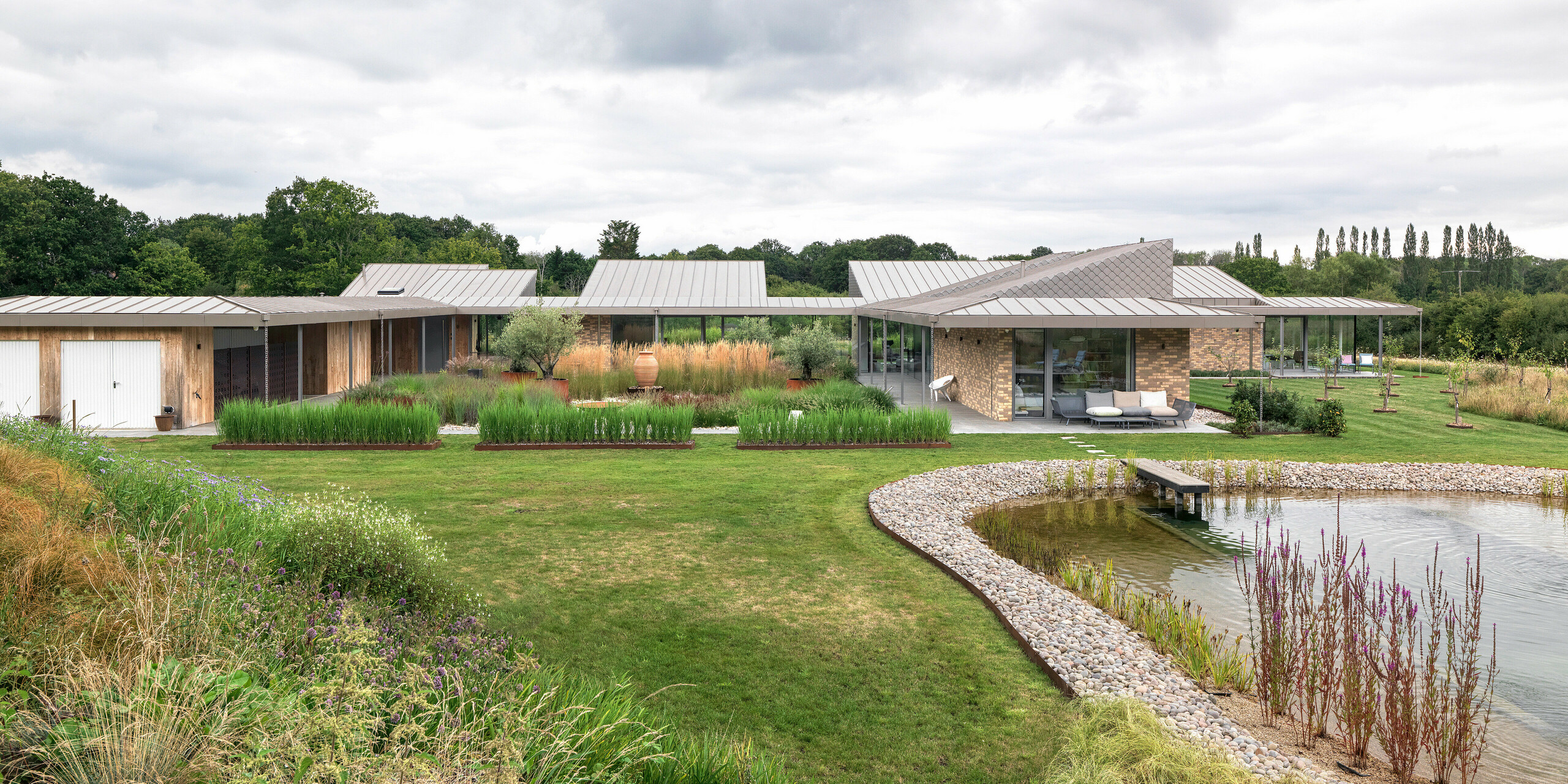 Wide-angle view of the architecturally sophisticated residential building in West Sussex with a large standing seam aluminium roof in P.10 bronze and harmoniously integrated PREFA rhomboid façade tiles in selected areas of the façade.The modern building extends flat and spacious across the property, nestled in natural landscaping with a landscaped natural pool in the foreground. The robust PREFA products support the sustainable building concept and ensure a uniform, long-lasting appearance.